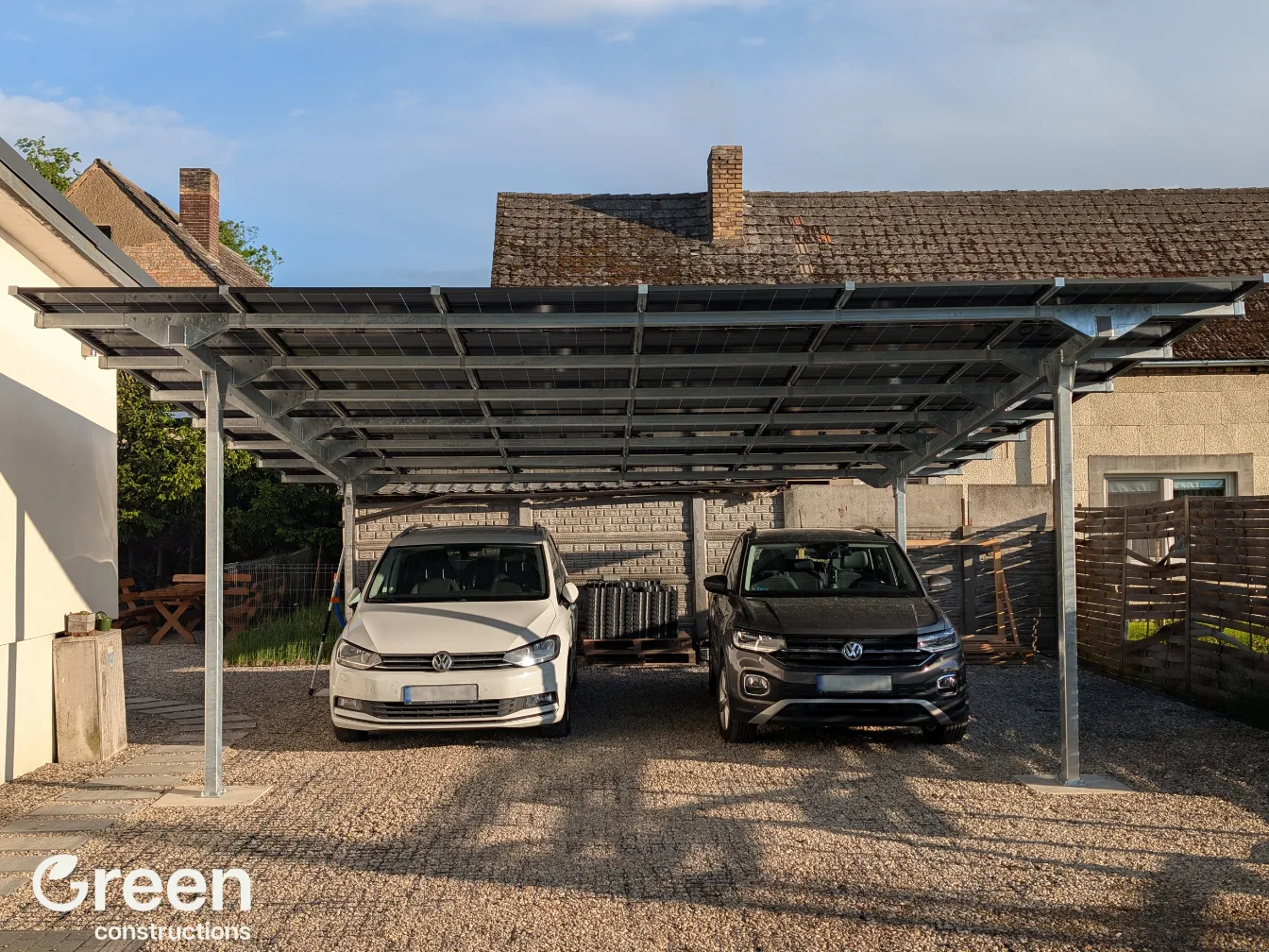 Wiata Fotowoltaiczna o mocy 12 kWp posadowiona na 4 słupach. Widok od strony wjazdu, pod wiata są zaparkowane 2 pojazdy. Na wiacie są zainstalowane 24 moduły fotowoltaiczne.