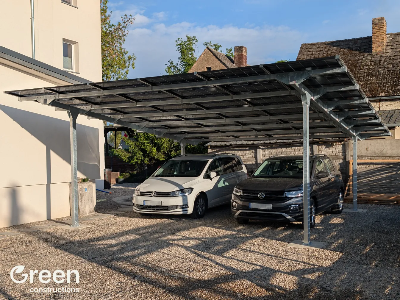 Wiata Fotowoltaiczna o mocy 12 kWp. Widok pod katem od strony wjazdu, pod wiata są zaparkowane 2 pojazdy, sam teren jest wykończony szarym kamyszkiem. Na wiacie są zainstalowane 24 moduły fotowoltaiczne.